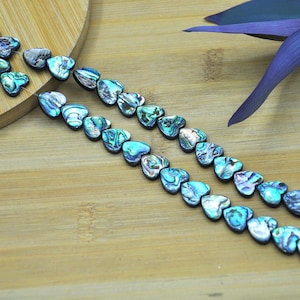 Peut inclure: Deux rangées de perles en forme de cœur aux tons de bleu, vert et argent, avec des motifs irisés. Les perles sont disposées sur une surface en bois brun clair, avec une feuille violette en arrière-plan. Les perles sont probablement destinées à la fabrication de bijoux.