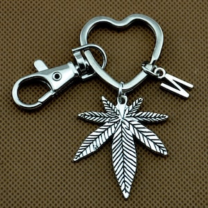 Op de afbeelding: Zilverkleurige sleutelhanger met een hartvormige ring, een karabijnhaak, een cannabisbladbedel en een letter "W" bedel. De cannabisbladbedel heeft gedetailleerde nerven.