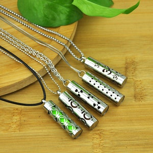 Op de afbeelding: Vijf zilveren kettingen met verschillende hangerontwerpen. Elke hanger is een holle cilinder met een ander uitgesneden patroon. De patronen omvatten een bloem, een cirkel, een stippenpatroon, een hart en een Om-symbol. De kettingen liggen op een houten oppervlak.