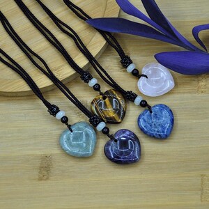 May include: Heart-shaped gemstone pendants on black cords. The pendants are in various colors, including blue, purple, green, tiger's eye, and pink. Each necklace has a decorative bead near the pendant.