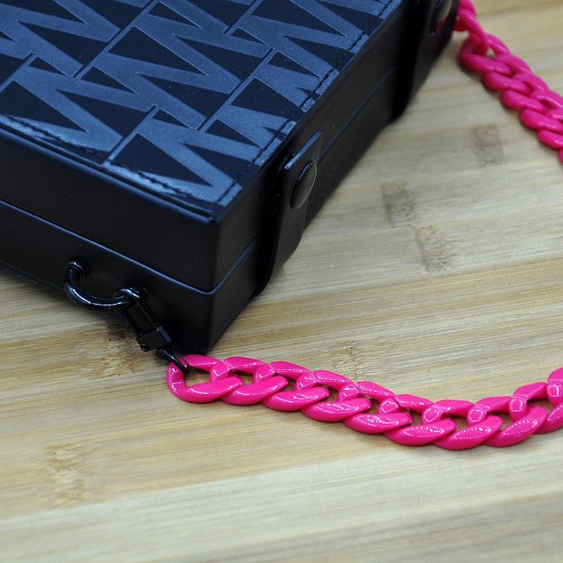 May include: A black box with a geometric pattern and a pink chain strap. The chain is made of thick, chunky links.