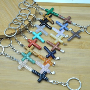 May include: A collection of cross-shaped keychains in various colors, including green, red, blue, and brown. Each cross is made of a different stone and attached to a silver-colored chain and keyring. The keychains are arranged on a wooden surface.
