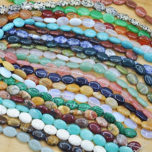 Puede incluir: Una colección de cuentas de piedras preciosas de forma ovalada en varios colores, incluyendo turquesa, rojo, verde y marrón. Las cuentas están ensartadas, mostrando una variedad de texturas y patrones. Las cuentas se exhiben sobre una superficie de madera clara.