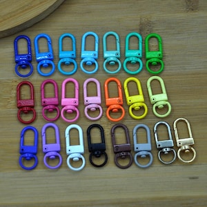 May include: Assortment of 20 colourful metal swivel lobster clasps. The clasps are in various colours, including red, orange, yellow, green, blue, purple, pink, and black. The clasps are all the same size and shape.