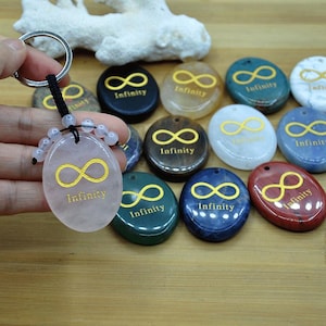 May include: A collection of oval-shaped stone pendants with the infinity symbol and the word "Infinity" in gold. One pendant is attached to a keychain. The pendants are in various colors, including pink, green, blue, and red.