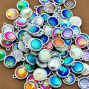 Peut inclure: Un grand tas de breloques écailles de sirène irisées avec des montures en métal argenté. Les breloques sont rondes et présentent une variété de couleurs, notamment le bleu, le vert, le rose et le violet.