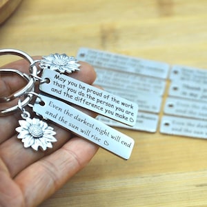 以下が含まれることがあります： 銀色のキーホルダーに2つのタグが付いています。 上のタグには「May you be proud of the work that you do. The person you are and the difference you make.」と書かれています。 下のタグには「Even the darkest night will end and the sun will rise.」と書かれています。