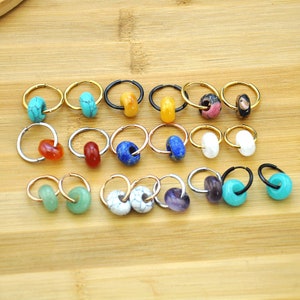 Pode incluir: Uma coleção de brincos de argola com contas coloridas. Os brincos apresentam argolas douradas, prateadas e pretas, cada uma adornada com contas em tons de turquesa, laranja, azul, branco e roxo. Os brincos são exibidos em uma superfície de madeira marrom clara.