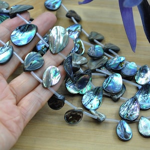 Op de afbeelding: Abalone schelpkralen in tinten blauw, groen en zilver zijn geregen op een transparante streng. De druppelvormige kralen worden gepresenteerd op een houten oppervlak.