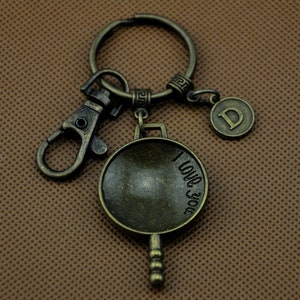 Puede incluir: Un llavero de color bronce con un pequeño dije en forma de sartén que dice "Te amo" y un dije con la letra "D". El llavero tiene un anillo de metal y un cierre de langosta.