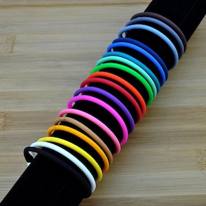 May include: A collection of fifteen thin, circular silicone bracelets arranged in a row on a black display stand. The bracelets feature a smooth, matte finish and come in a vibrant spectrum of colors including black, white, yellow, orange, pink, purple, blue, green, and brown.