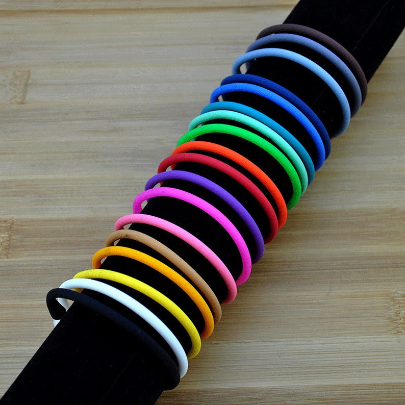 May include: A collection of fifteen thin, circular silicone bracelets arranged in a row on a black display stand. The bracelets feature a smooth, matte finish and come in a vibrant spectrum of colors including black, white, yellow, orange, pink, purple, blue, green, and brown.