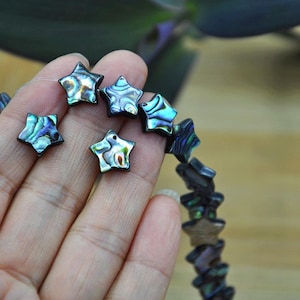 May include: Star-shaped beads made from abalone shell, displaying iridescent hues of blue, green, and purple. The beads are strung together, with a few loose ones visible. The beads are held in a hand, highlighting their natural patterns and colours.