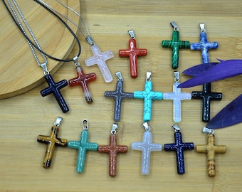 May include: A selection of gemstone cross pendants on necklaces. The crosses are in various colours and materials, including red, blue, green, and brown. Each pendant has a silver-coloured bail. The necklaces are displayed on a wooden surface.