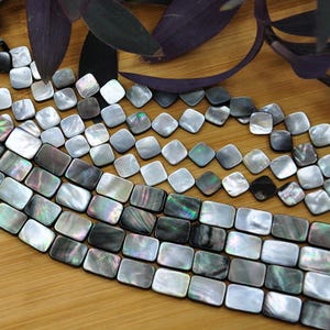 May include: A collection of iridescent mother-of-pearl beads in square and rectangular shapes. The beads feature a smooth, polished finish with a natural colour palette ranging from light grey and silver to deep charcoal, displaying a shimmering rainbow lustre.