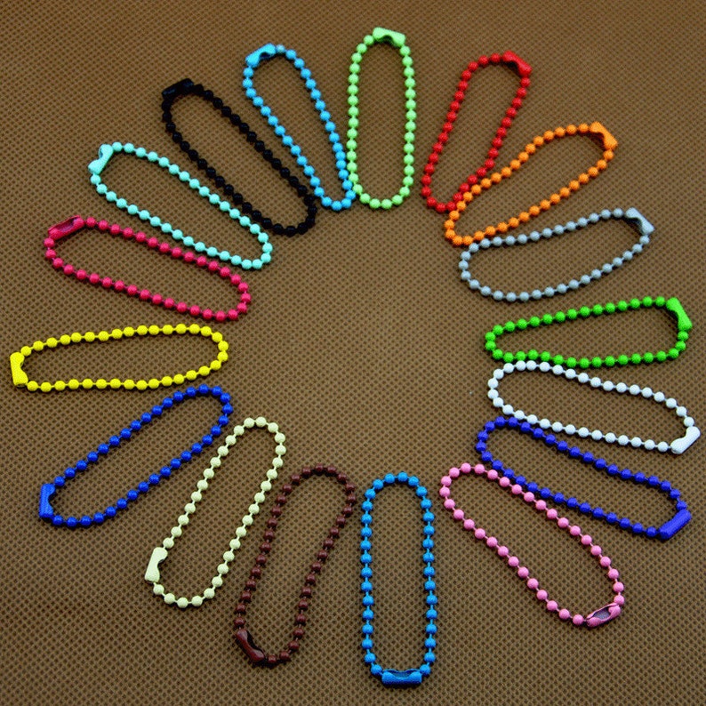 Pode incluir: Um conjunto de 20 chaveiros de metal coloridos, cada um com uma corrente de pequenas contas redondas. Os chaveiros est&atilde;o dispostos em c&iacute;rculo sobre uma superf&iacute;cie marrom texturizada.