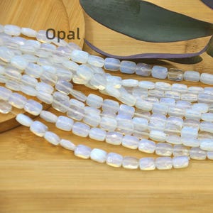 Puede incluir: Hebras de cuentas de ópalo iridiscentes, de corte cuadrado. Las cuentas son de color blanco lechoso con sutiles tonos azules y morados. La palabra "Opal" está impresa en negro sobre una superficie de madera.