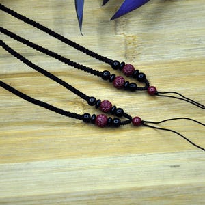 May include: Close-up of three black cord necklaces with decorative beads. Each necklace features a series of small black beads, a cluster of textured red beads, and a single red bead at the end. The necklaces are displayed on a light brown wooden surface.