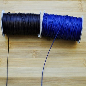 May include: Two spools of waxed cord, one black and one blue, are shown on a wooden surface. The cords are unwound and hanging down.