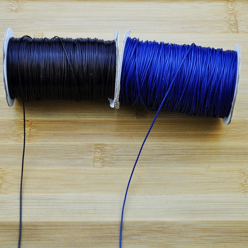 May include: Two spools of waxed cord, one black and one blue, are shown on a wooden surface. The cords are unwound and hanging down.