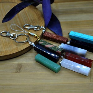 May include: A selection of gemstone keyrings in diverse colours and patterns, including green, red, blue, and speckled designs. Each keyring features a rectangular gemstone pendant attached to a silver-tone clasp and ring. The keyrings are displayed on a wooden surface.