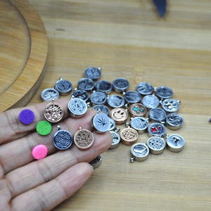 May include: A collection of small, round pendants with various designs, including a smiley face, a leaf, and floral patterns. The pendants are silver and bronze-toned, with a few colorful accents. Also included are three small, colorful felt pads.