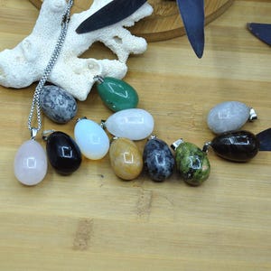 May include: A collection of gemstone egg pendants in various colors, including pink, black, green, and white. Each pendant has a silver-tone bail and is displayed on a wooden surface. Some pendants are on a silver chain.