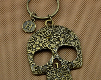 May include: A bronze-colored key chain with a skull charm. The skull is decorated with a floral pattern and the letter "H" is on a smaller charm attached to the key ring.
