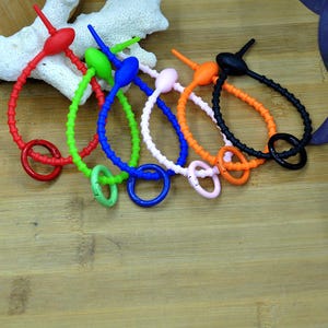 May include: A collection of six flexible silicone wristbands in red, green, blue, light pink, orange, and black. Each band features a textured, beaded surface, a small oval-shaped toggle, and a matching circular ring clasp attached at the end.