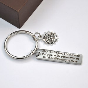 Könnte beinhalten: Silberner Schlüsselanhänger mit einem Sonnenblumen-Anhänger und einem rechteckigen Anhänger mit der Aufschrift: "Möge Sie stolz auf die Arbeit sein, die Sie leisten, die Person, die Sie sind, und den Unterschied, den Sie machen."