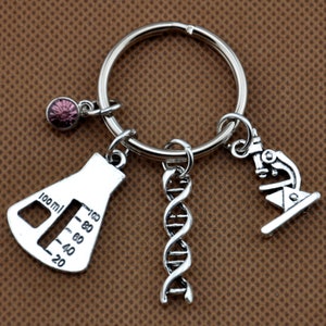 May include: Silver keychain with four charms: a beaker with a 100 ml measurement, a double helix DNA strand, a microscope, and a small pink rhinestone.