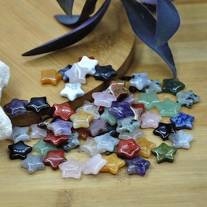 May include: A collection of small, star-shaped gemstones in various colours, including red, green, blue, and yellow. The gemstones are scattered on a wooden surface, with some purple leaves in the background. The stars are approximately 2 cm in size.