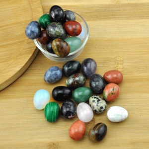 May include: A collection of tumbled gemstone eggs in various colours, including green, red, blue, black, and white. The eggs are arranged in a bowl and on a wooden surface.