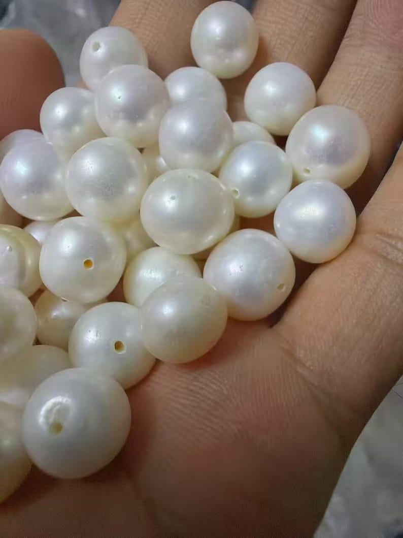 May include: A collection of loose, round, white pearls with a smooth, lustrous finish. Each individual pearl features a small, centered drill hole, indicating they are intended for use as beads in jewelry making or other decorative craft projects.