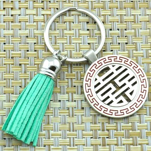 Puede incluir: Un llavero plateado con un borla verde y un dije redondo plateado con un borde rojo y un símbolo chino en su interior.