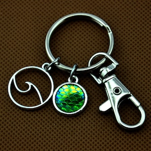 May include: Silver-coloured keyring with three charms. One charm is a circle with a wave design, another is a round charm with a green and yellow scale pattern, and the third is a lobster clasp.