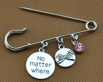 Pode incluir: Alfinete de segurança prateado com três pingentes. Um diz "No matter where", outro mostra mãos entrelaçadas, e o terceiro é um cristal rosa. Um item de amizade ou lembrança.