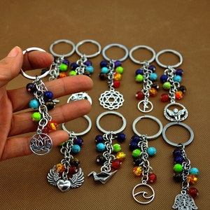 Puede incluir: Un conjunto de 10 llaveros de cuentas coloridas con diferentes dijes de plata. Los dijes incluyen una flor de loto, un corazón con alas, un pájaro, una ola y una mano de hamsa. Las cuentas son en tonos de rojo, naranja, amarillo, verde, azul y morado.