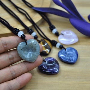 May include: Heart-shaped gemstone pendants on black cords. The pendants are various colors, including gray, tiger's eye, pink, blue, and purple. Each pendant is unique, with variations in color and texture. The cords have decorative beads.