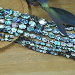 Può includere: Una collezione di perline iridescenti di conchiglia di abalone in varie dimensioni e forme. Le perline mostrano una gamma di colori, tra cui blu, verdi e viola, con un effetto scintillante e perlaceo. Le perline sono disposte su una superficie di legno.