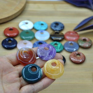 以下が含まれることがあります： 中央に穴の開いた、カラフルな丸い宝石ペンダントのコレクション。ペンダントは赤、緑、青、紫、黄色など様々な色があります。宝石は滑らかで磨かれており、光沢のある仕上がりです。