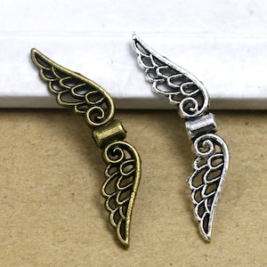 May include: Two metal wing charms. One is bronze-colored, the other is silver. Both have a detailed, openwork design with curved lines and a connecting bar. These charms are suitable for jewelry making or craft projects.