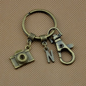 Könnte beinhalten: Ein bronzefarbener Schlüsselanhänger mit einem Kamera-Anhänger, dem Buchstaben "N" und einem Verschluss. Der Kamera-Anhänger hat ein detailliertes Design, und der Schlüsselanhänger ist an einem kreisförmigen Ring befestigt. Der Hintergrund ist einfarbig braun.