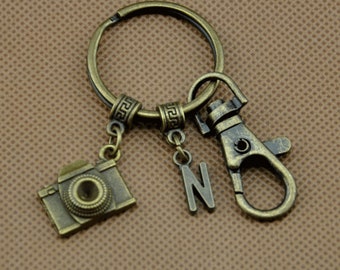 May include: A bronze-toned keyring with a camera charm, the letter "N", and a clasp. The camera charm has a detailed design, and the keyring is attached to a circular ring. The background is a solid brown colour.