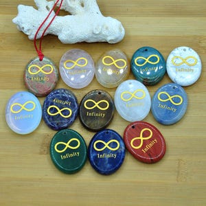 Peut inclure: Une collection de pendentifs ovales de différentes couleurs, chacun avec un symbole infini doré et le mot "Infinity". Les pendentifs sont faits de différentes pierres et sont présentés sur une surface en bois.