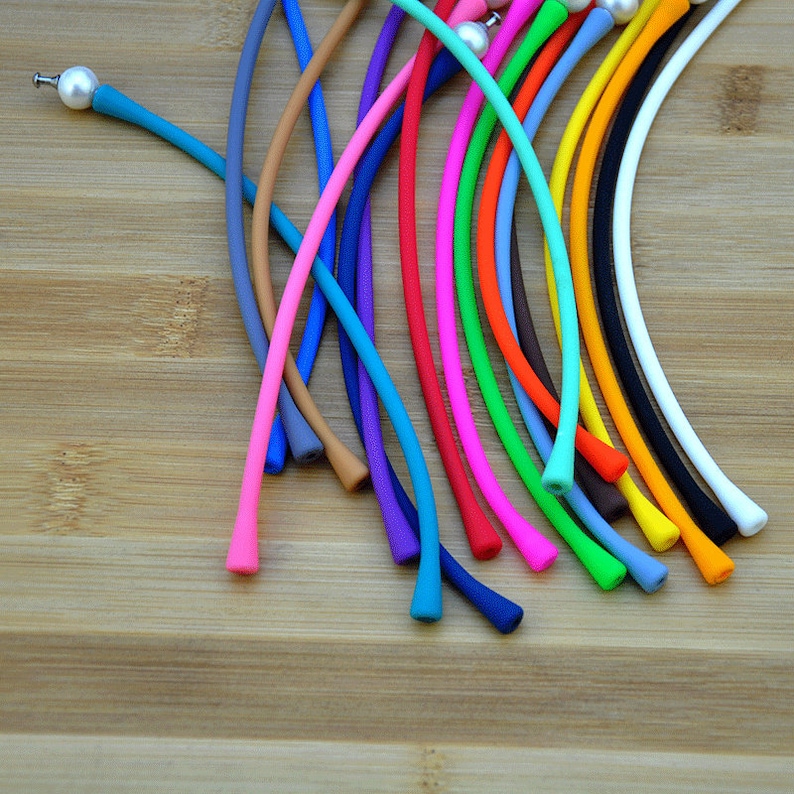May include: A collection of flexible, smooth silicone cords in a variety of vibrant colors including blue, pink, orange, green, yellow, black, and white. Each cord features a small, round, white pearl-like bead attached to one end.