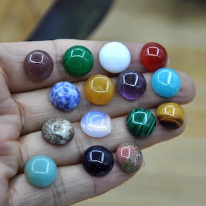 May include: A selection of polished gemstone cabochons in various colours, including green, blue, red, and yellow. The stones are round and smooth, with a glossy finish. The gemstones are held in a hand.