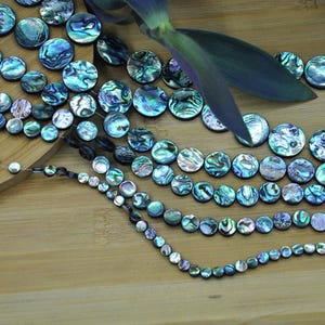 Könnte beinhalten: Eine Sammlung schillernder Abalone-Muschelkernperlen in verschiedenen Größen und auf zarten Strängen aufgereiht. Die Perlen zeigen eine Reihe von Blau-, Grün- und Lilatönen mit einem natürlichen, strukturierten Aussehen. Einige Perlen sind lose, andere in Reihen angeordnet.