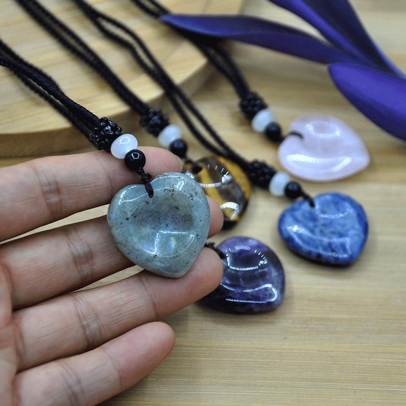 May include: Heart-shaped gemstone pendants on black cords. The pendants are various colors, including blue, pink, and green. The gemstones are smooth and polished, with a natural, organic look. The cords have white and black beads.