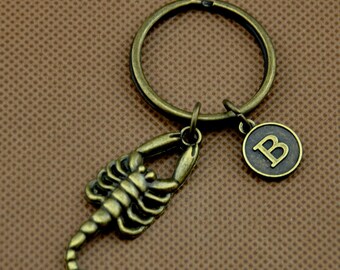 Puede incluir: Un llavero de color bronce con un dije de escorpión y un dije de la letra 'B'. El llavero está unido a un anillo de llave plateado.